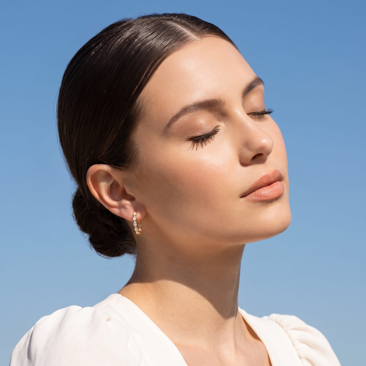 Woman wearing the Albane teardrop hoop gold earrings on the ear with eyes closed outdoors