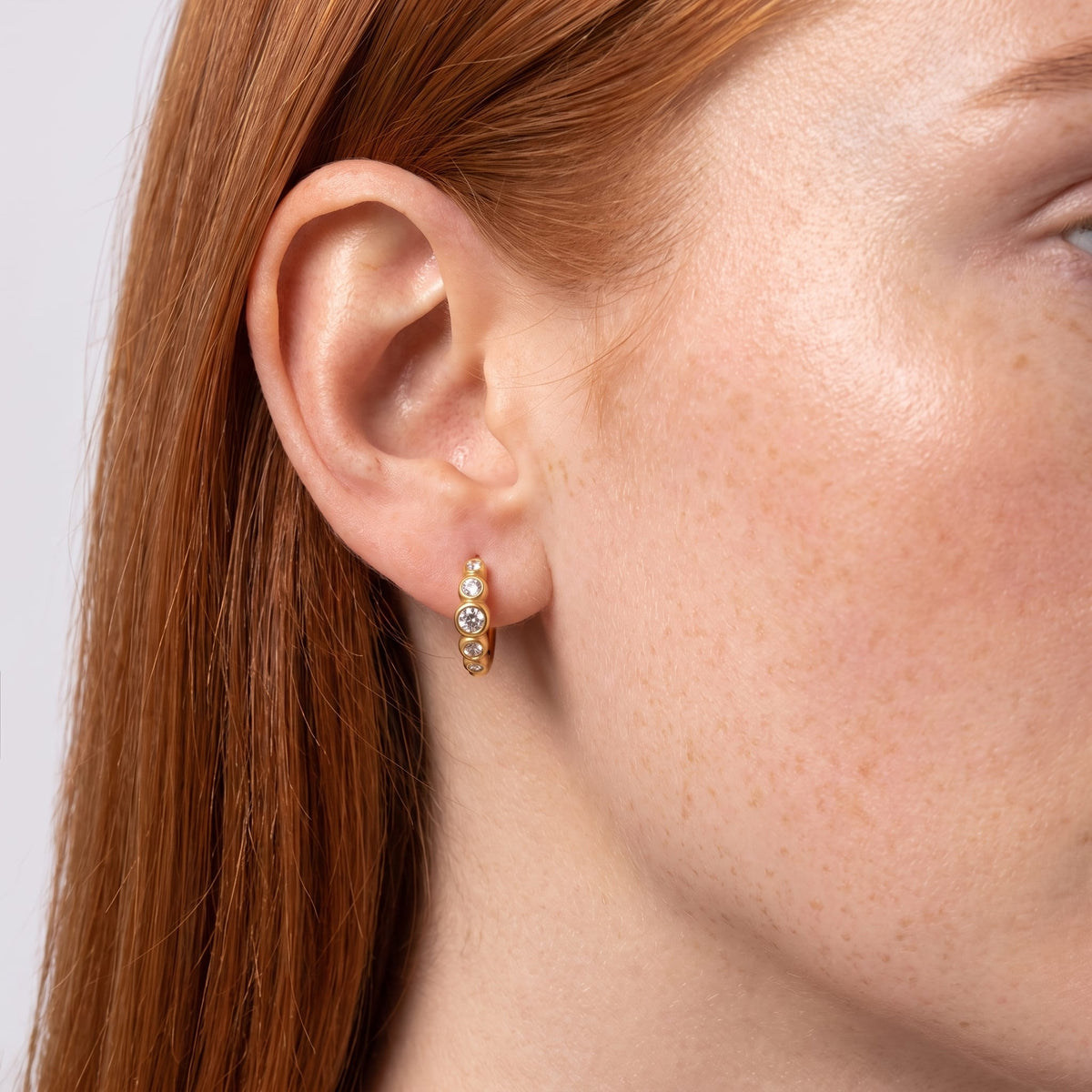 Woman wearing the Anaïs crystal huggie gold earrings on her earlobe with her red hair tucked behind