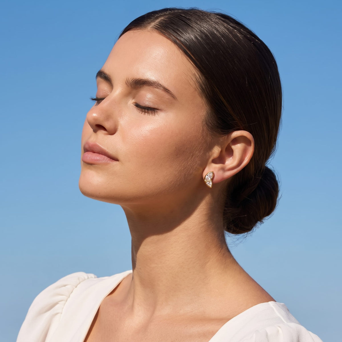 Woman wearing the Aurore crystal wing gold earrings on her ear with eyes closed against blue sky