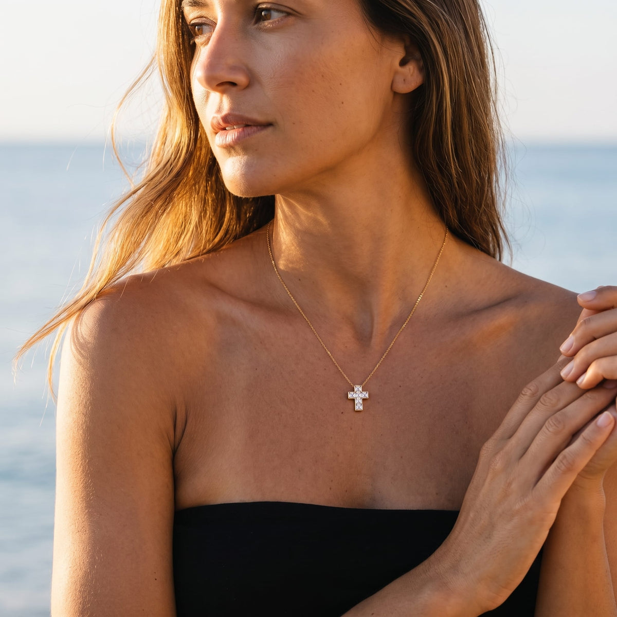 Woman wearing the Chiara crystal cross gold necklace at the collarbone near the ocean
