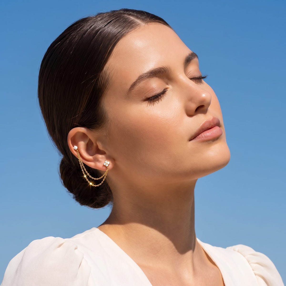Woman wearing the Félicie flower chain gold earrings on her ear against a clear blue sky