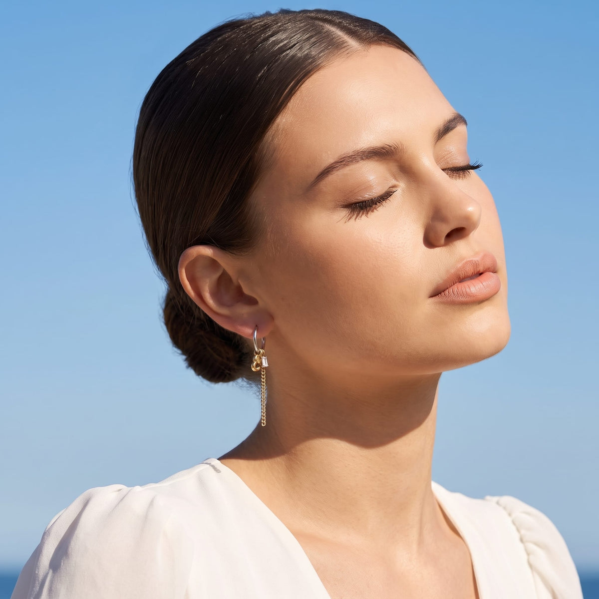 Woman wearing the Greta heart chain two-tone earrings on the ear with eyes closed in sunlight