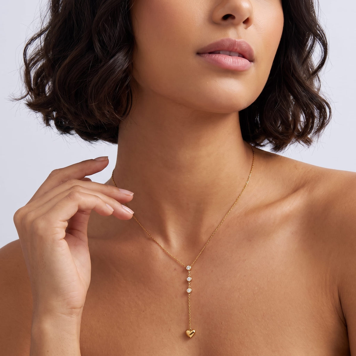 Woman wearing the Ilaria heart drop gold necklace at the collarbone while resting her hand near her neck