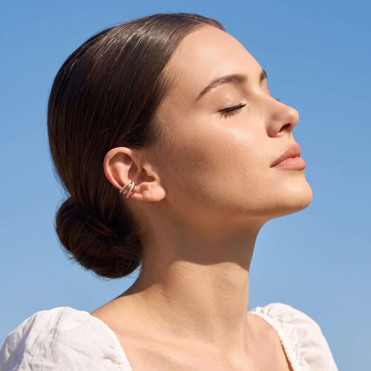 Woman wearing the Jeanne crisscross gold ear cuffs on the ear under bright blue sky