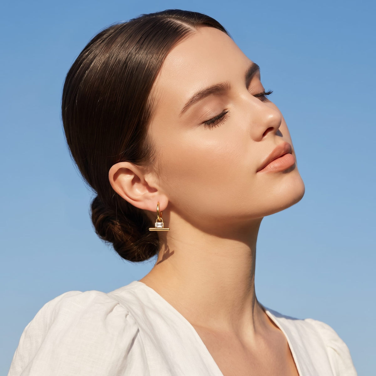 Woman wearing the Margaux padlock t-bar gold earrings on her ear against a bright blue sky