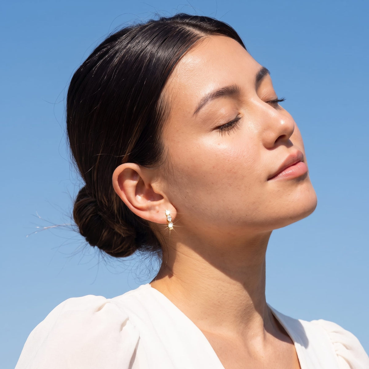 Woman wearing the Zoé marquise huggie gold earrings on the ear with eyes closed under a blue sky