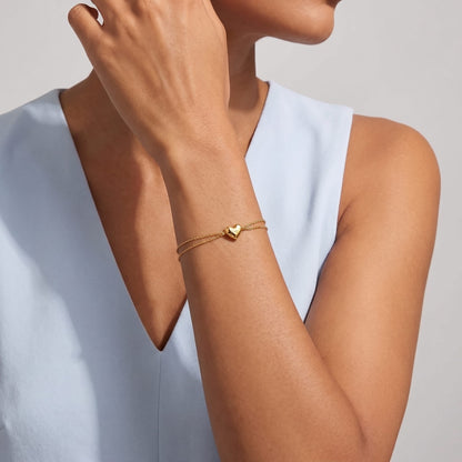 Woman wearing the Adèle hammered heart gold bracelet on the wrist wearing a light blue V-neck top