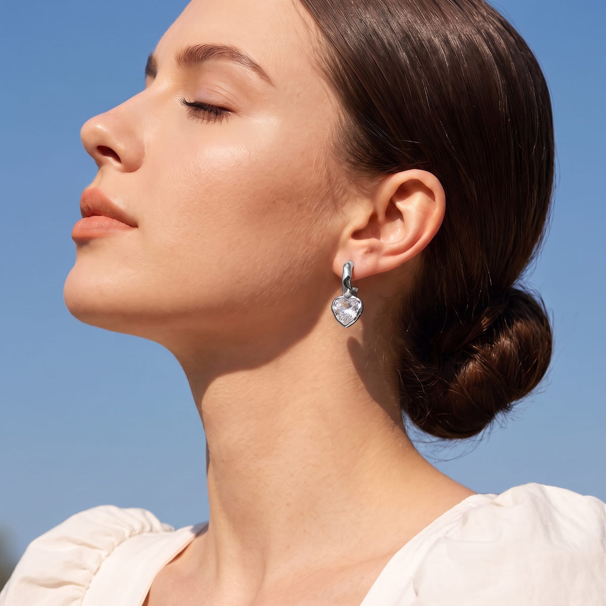 Woman wearing the Amandine hammered heart silver earrings on her ear with eyes closed against blue sky