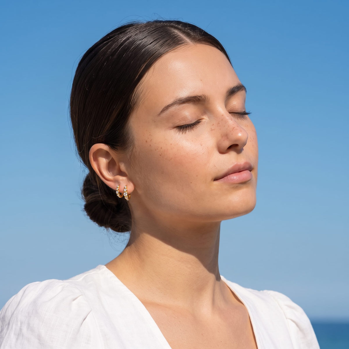 Woman wearing the Anaïs crystal huggie gold earrings on her earlobe against a clear blue sky background