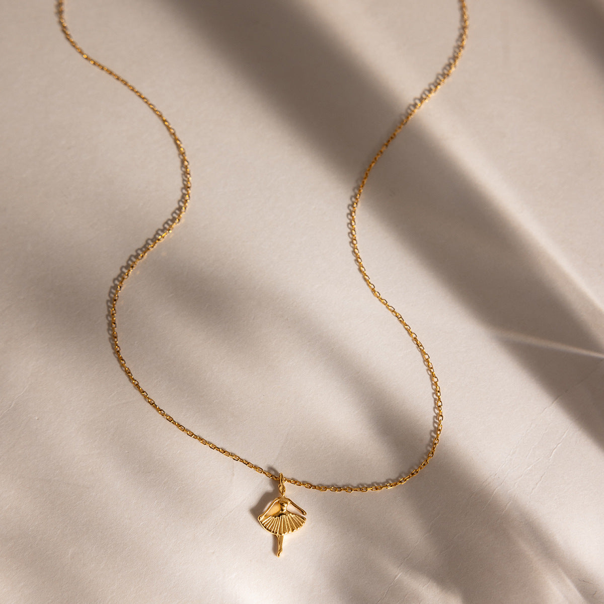 Ava ballerina necklace curving across pale linen in streaks of window shadow