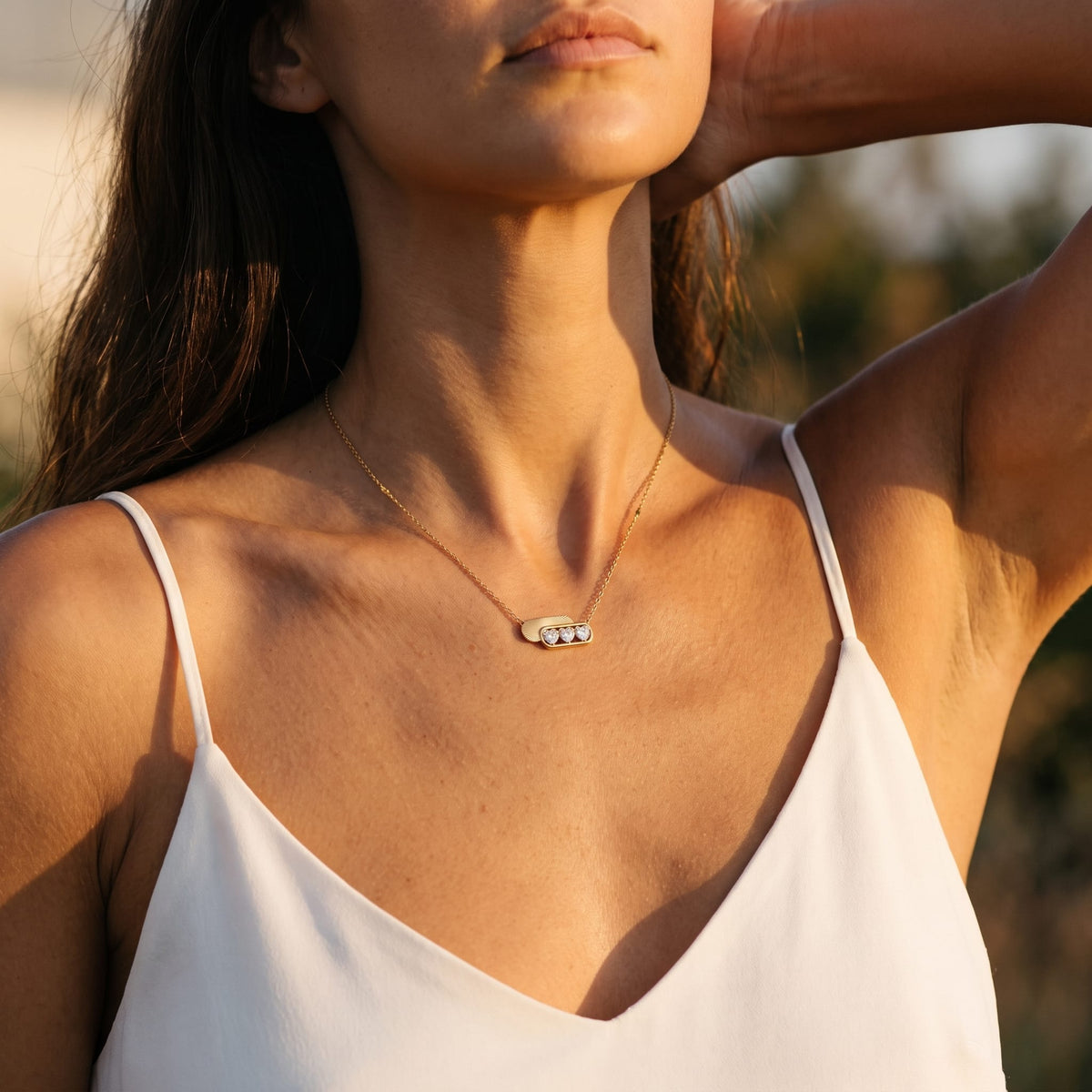 Woman wearing the Ginevra triple heart gold necklace at the collarbone raising her arm in golden hour light