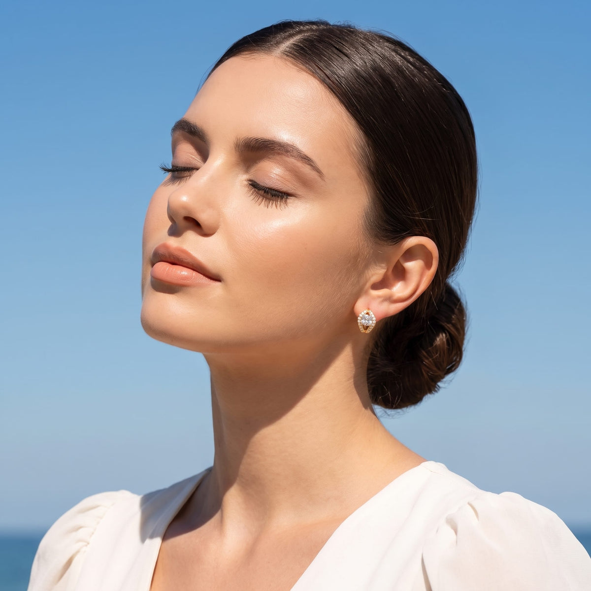 Woman wearing the Ilaria cushion halo gold earrings on the ear with eyes closed in sunlight