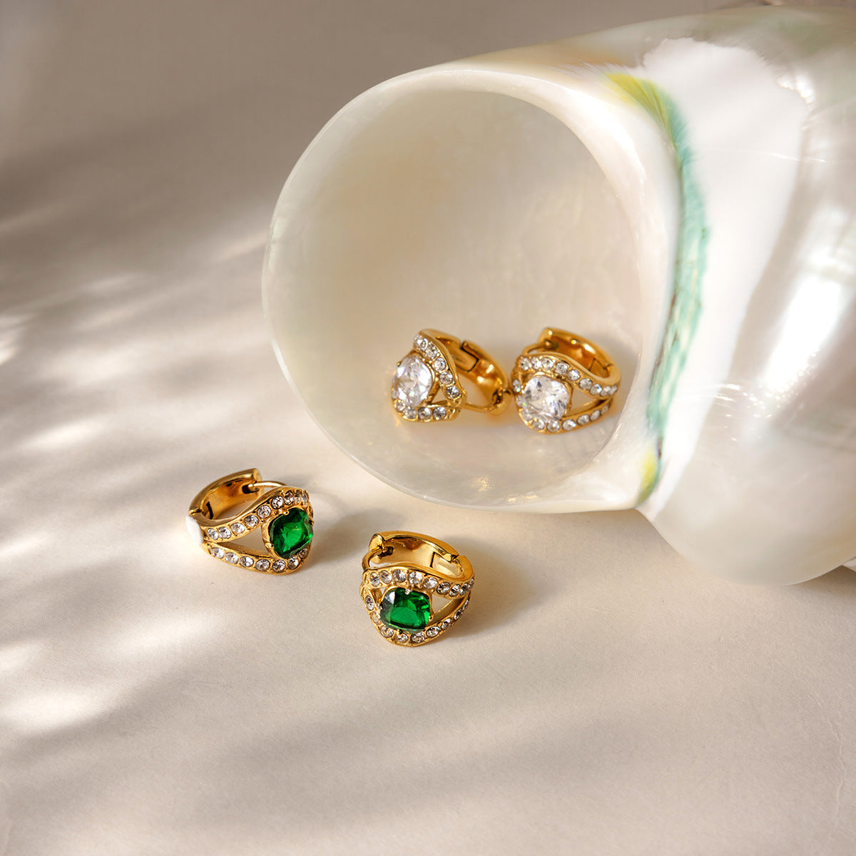 Green Ilaria huggies on a white surface beside a pearly shell holding the clear pair