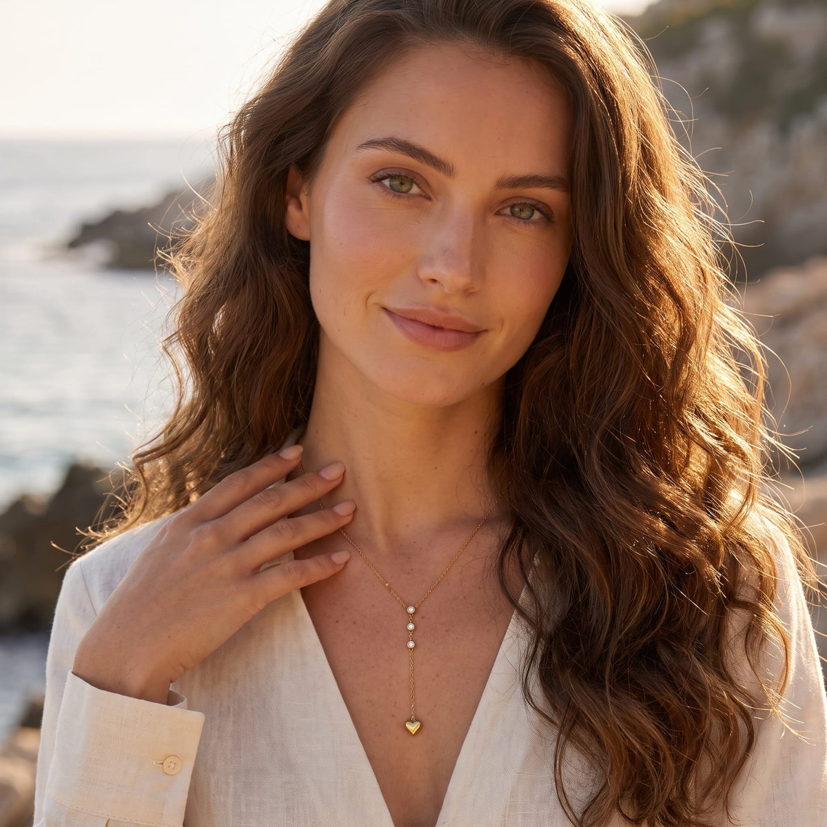 Woman wearing the Ilaria heart drop gold necklace at the collarbone against an outdoor coastal background