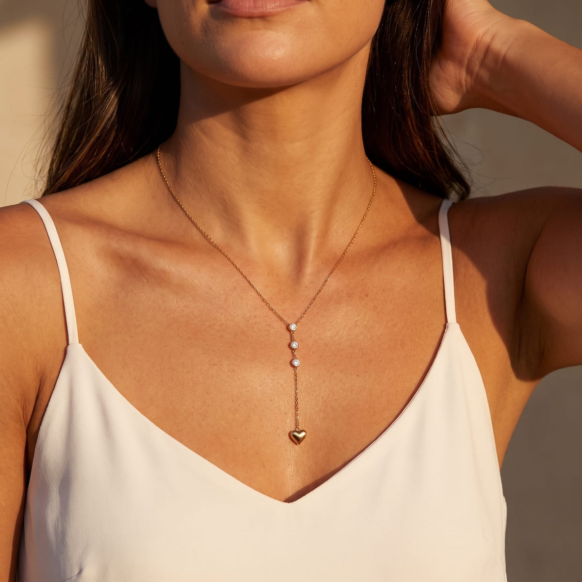 Woman wearing the Ilaria heart drop gold necklace at the collarbone in warm sunlight wearing a white camisole