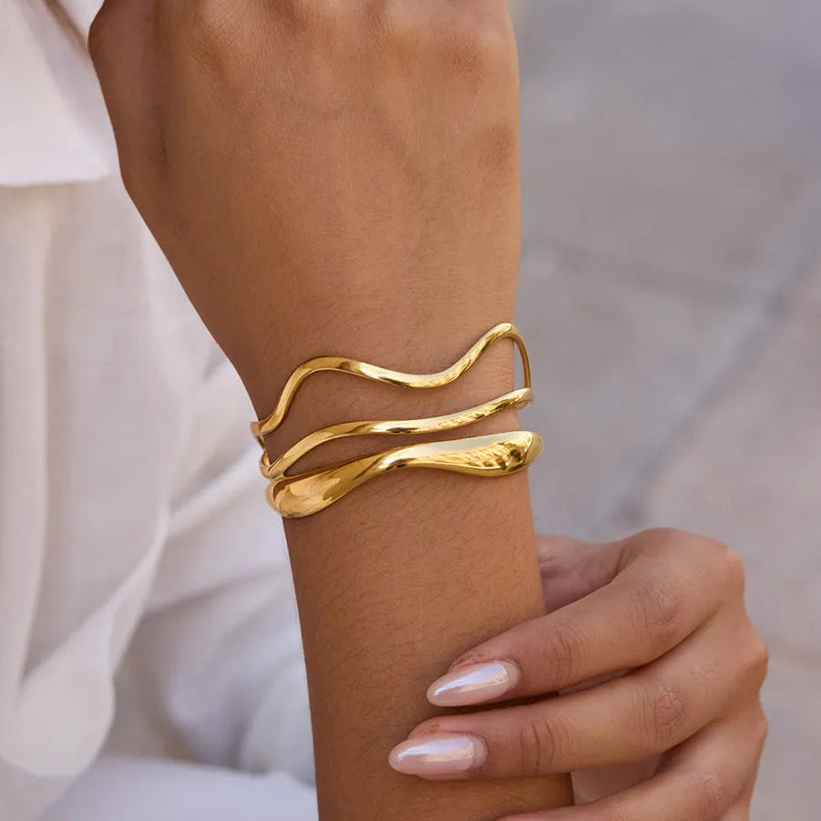 Three Emma wave gold bangles stacked on the wrist beside a white shirt sleeve