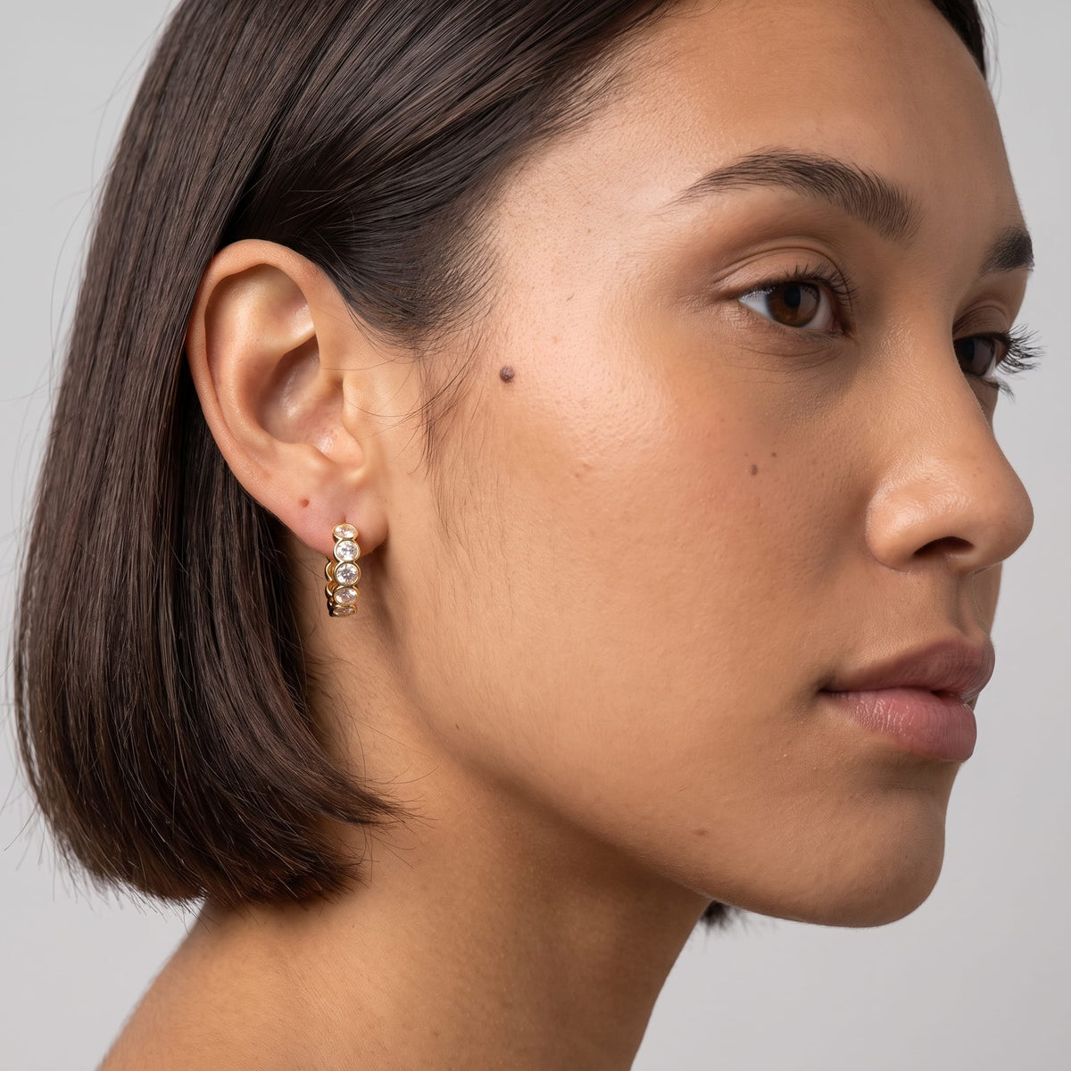 Woman wearing the Léa bezel huggie gold earrings on the ear in profile against a neutral backdrop