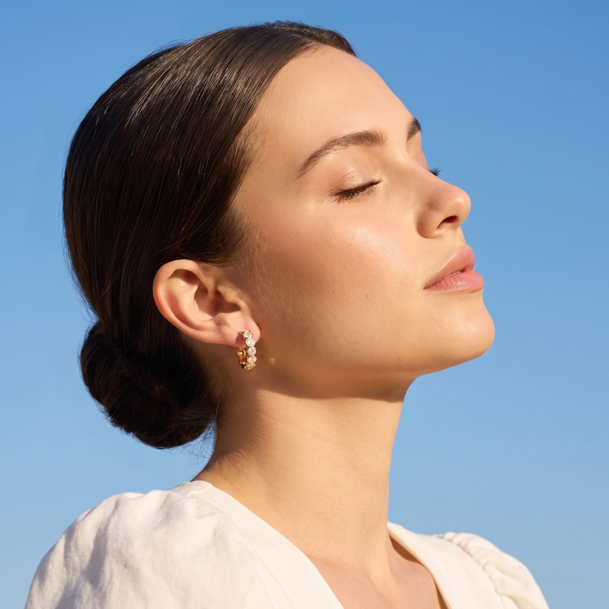 Woman wearing the Léa bezel huggie gold earrings on the ear facing up toward a blue sky
