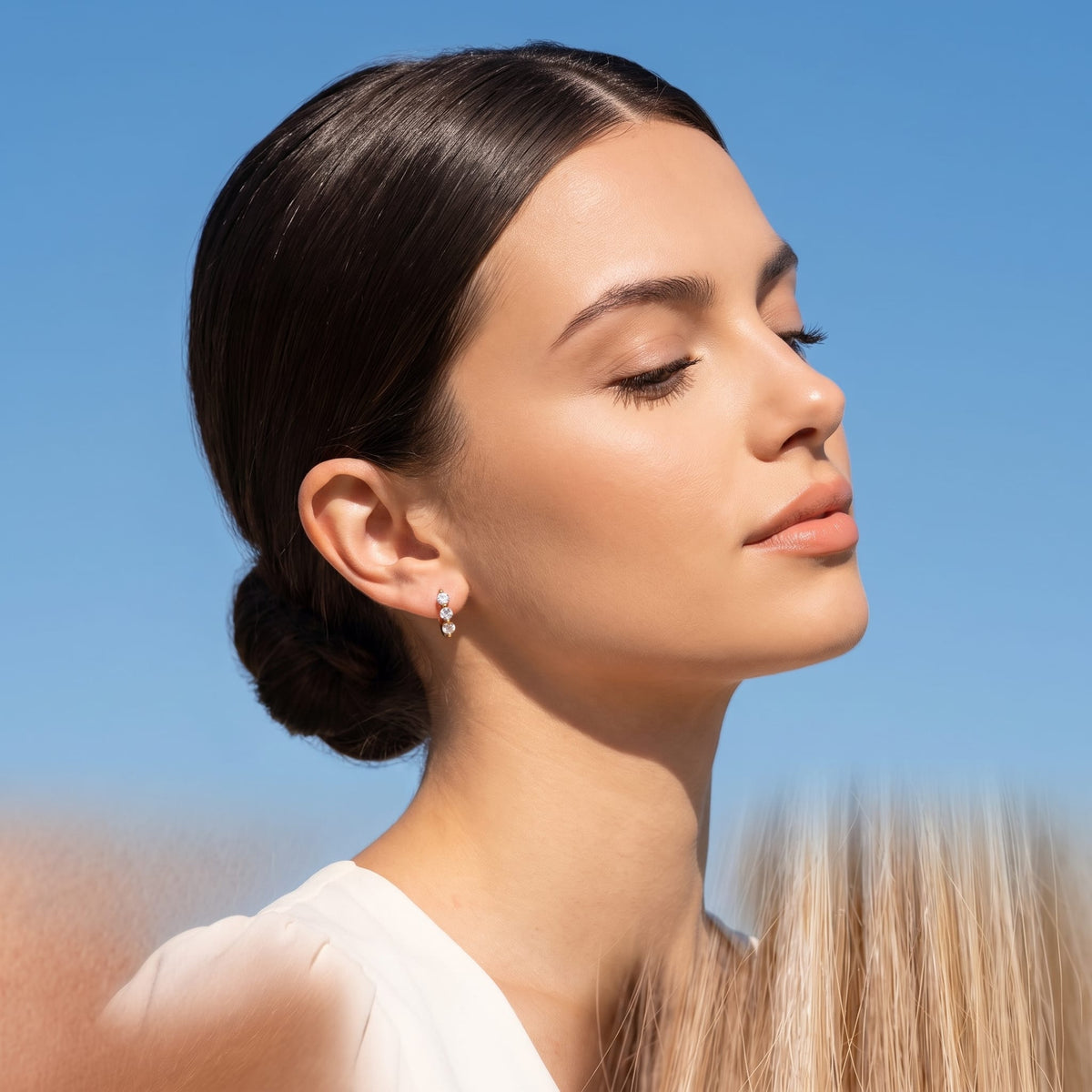 Woman wearing the Lucie three-stone huggie gold earrings on the ear against a blue sky