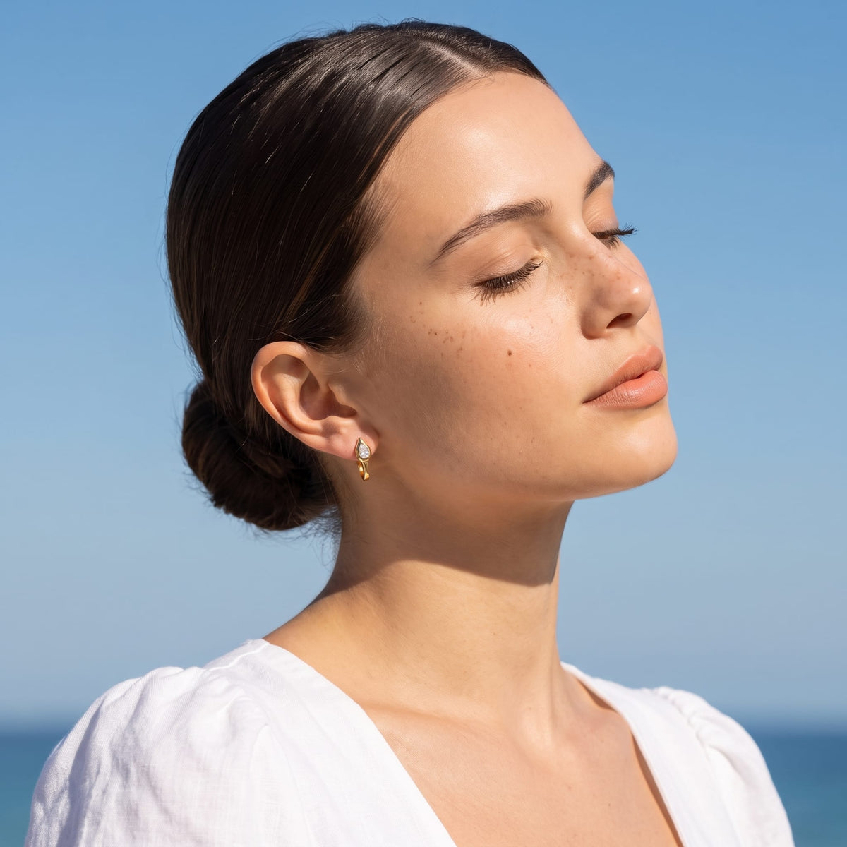 Woman wearing the Mireille pear bezel gold earrings on the ear wearing a white top outside