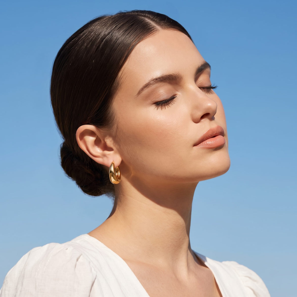 Woman wearing the Nina dome huggie gold earrings on the ear with a sleek bun against blue sky