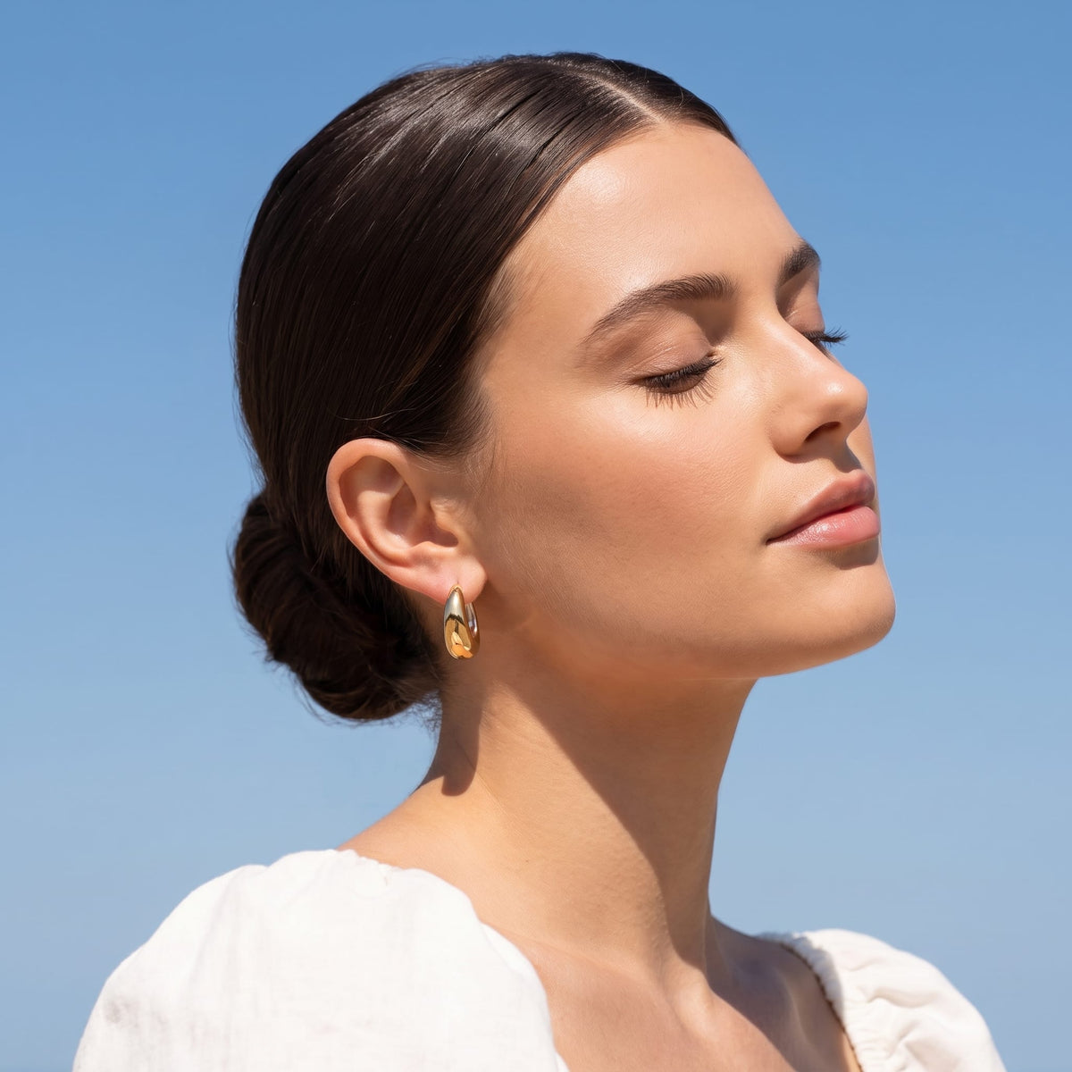 Woman wearing the Nina dome huggie two-tone earrings on the ear against a blue sky