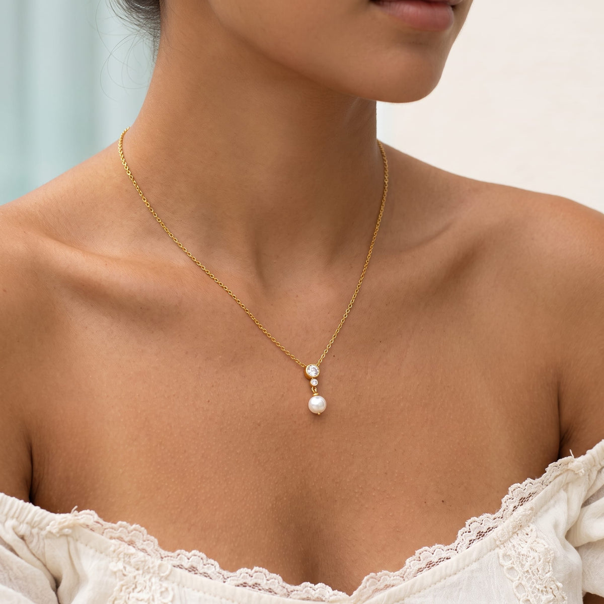 Wider view of a woman wearing the Odette crystal pearl gold necklace at the collarbone in a white lace top