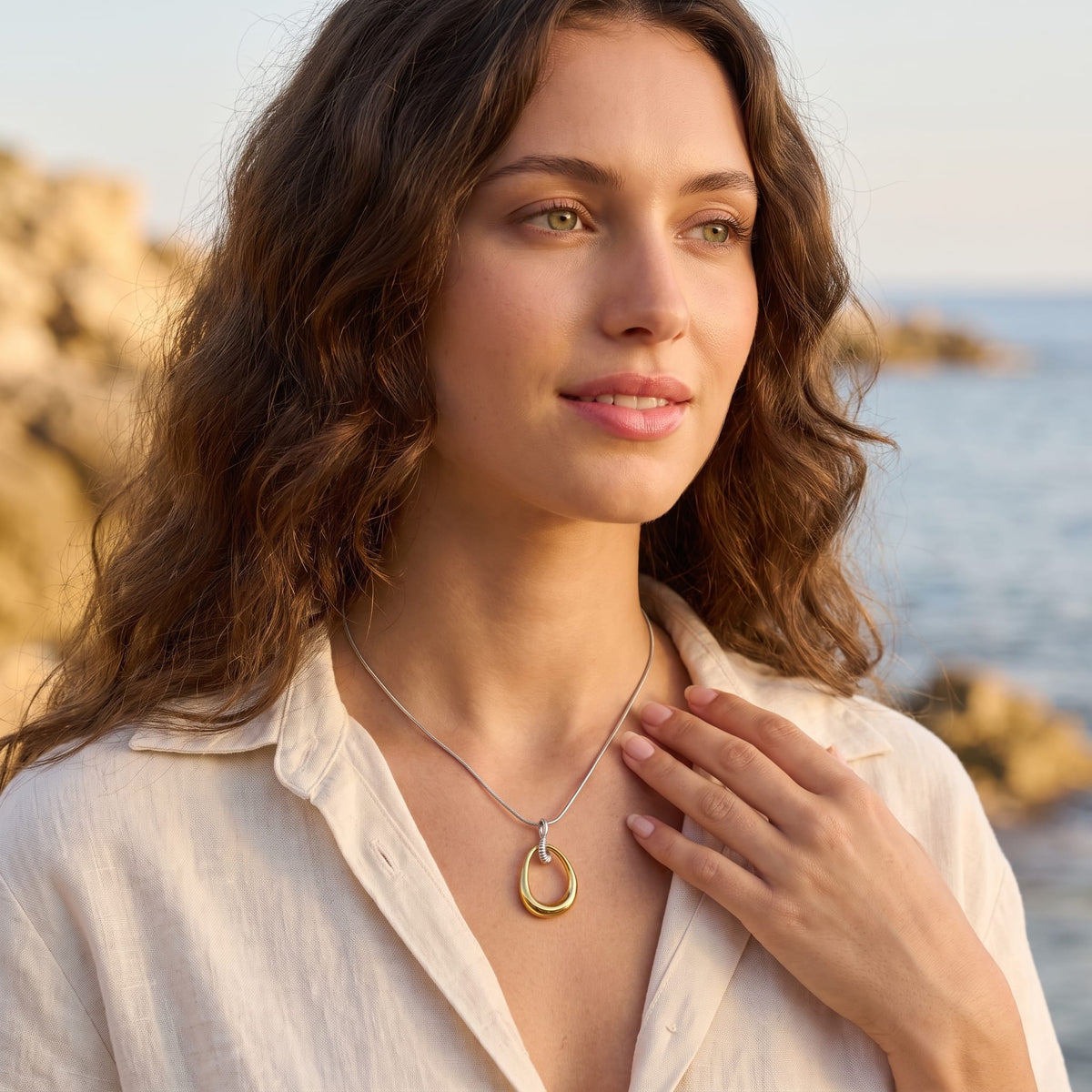 Woman wearing the Oriane open teardrop two-tone necklace at the collarbone near a sunny coastline