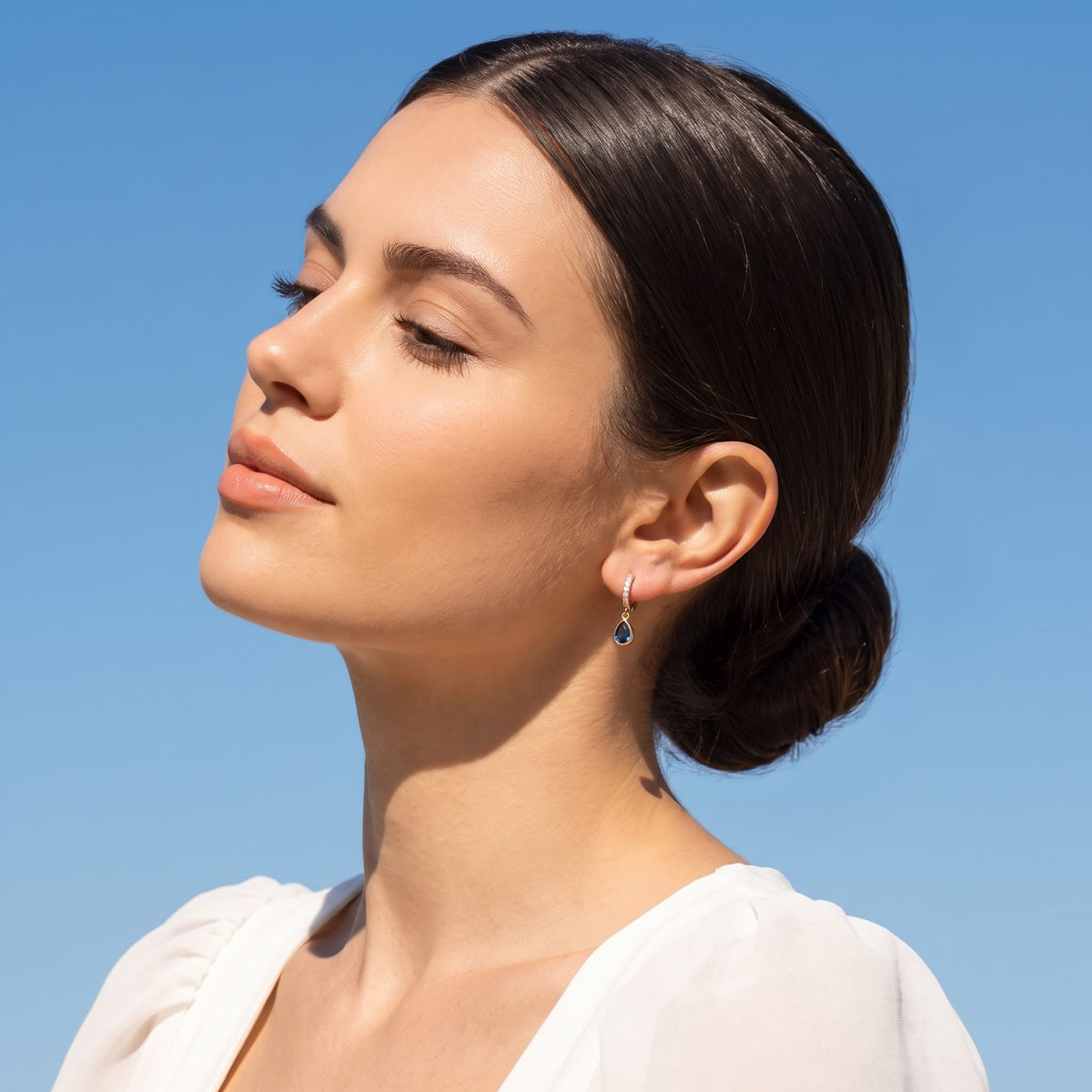 Woman wearing the Paola blue teardrop gold earrings on the ear against a clear blue sky