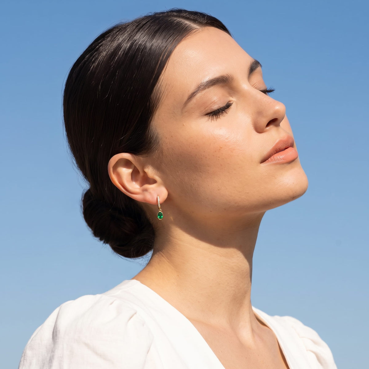 Woman wearing the Paola green oval gold earrings on the ear against a blue sky