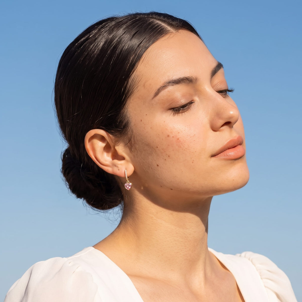 Woman wearing the Paola pink heart gold earrings on the ear wearing a white top with eyes closed