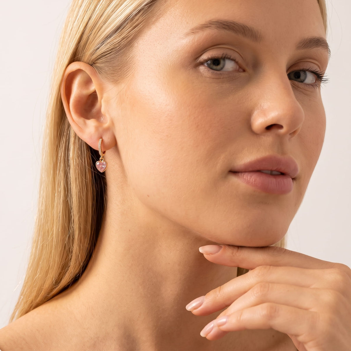 Woman wearing the Paola pink heart gold earrings on the ear resting her chin on her hand