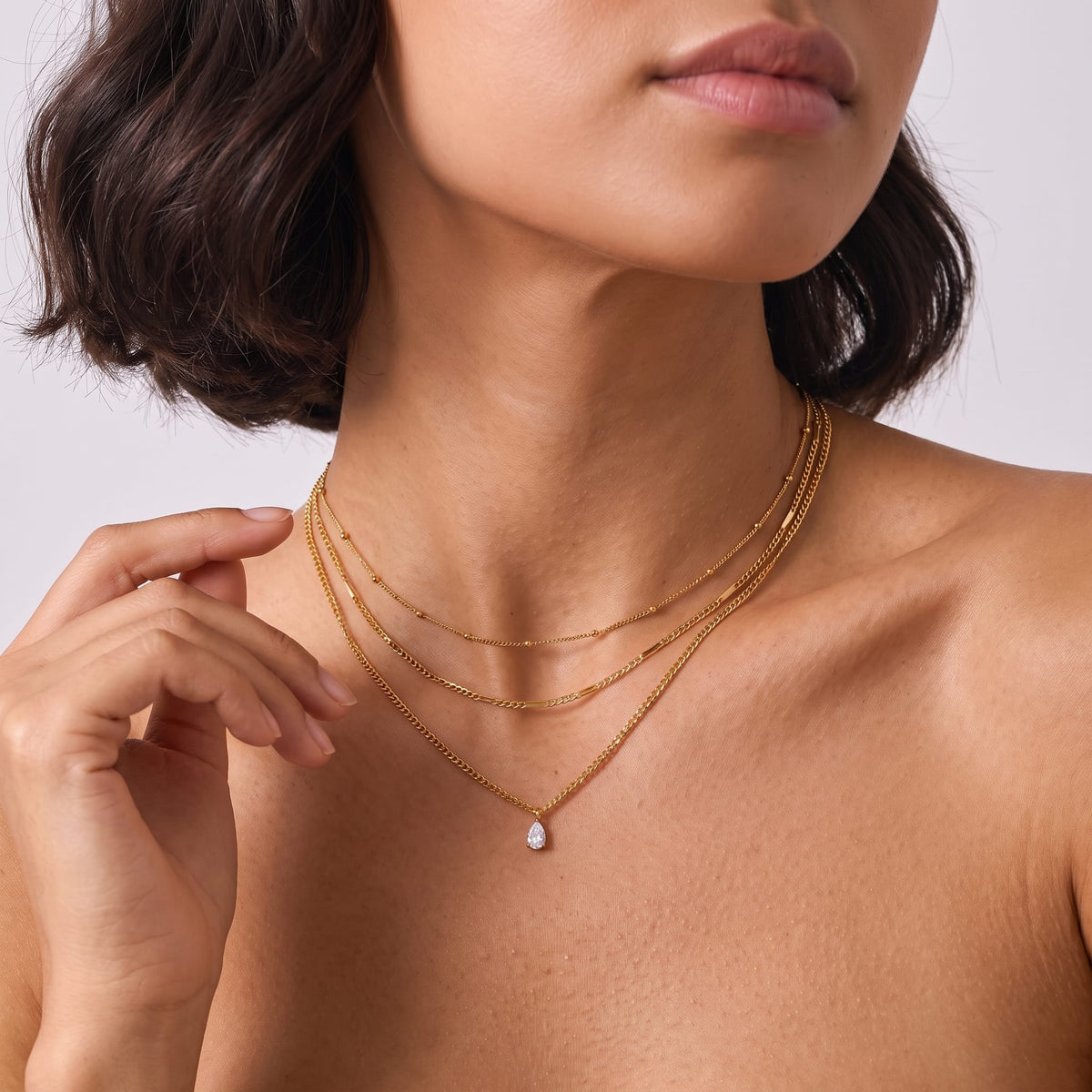 Woman wearing the Raphaëlle layered teardrop gold necklace at the throat, touching her neck against a plain backdrop