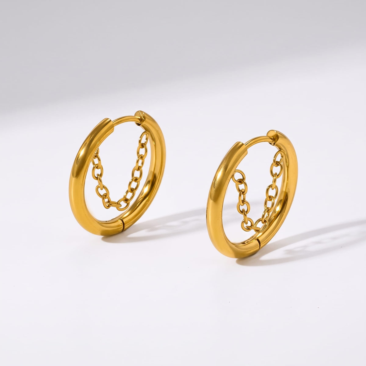 Romy chain hoop gold earrings on a plain white surface casting subtle soft shadows