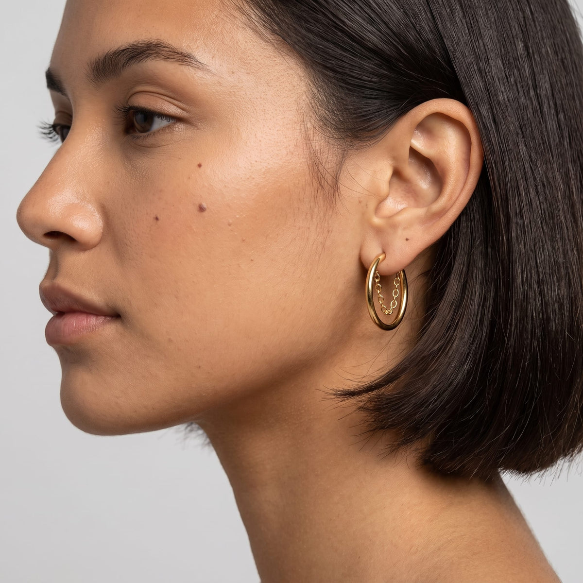 Woman wearing the Romy chain hoop gold earrings on the ear, in profile with short dark hair