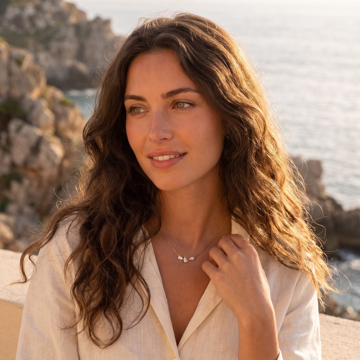 Woman wearing the Rosalie pear cluster gold necklace at the collarbone against a coastal backdrop