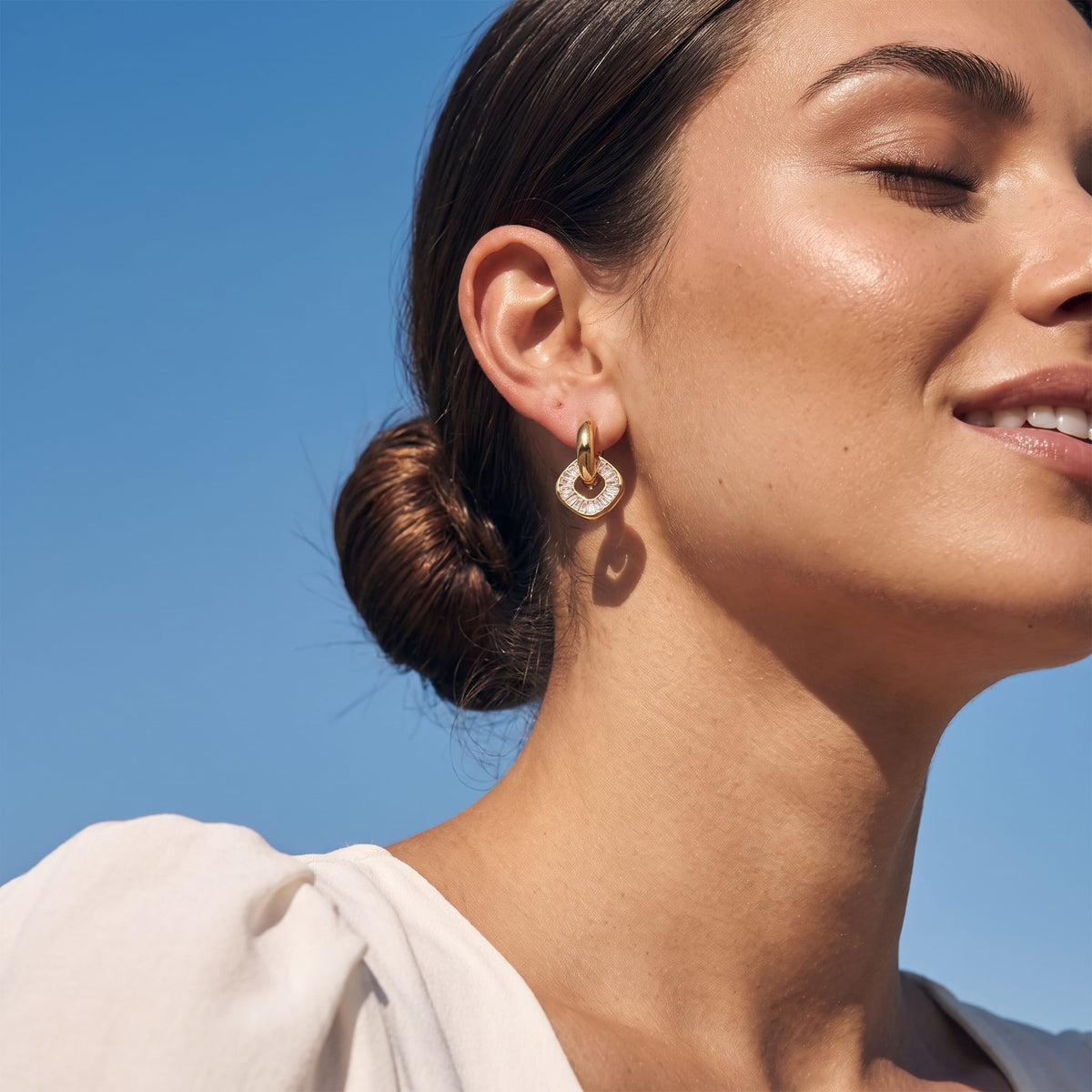 Woman wearing the Salomé baguette charm gold earrings on her ear under a blue sky